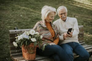 Senior couple sitting on the benchwith basket full of flowers and lookin at mobile phone